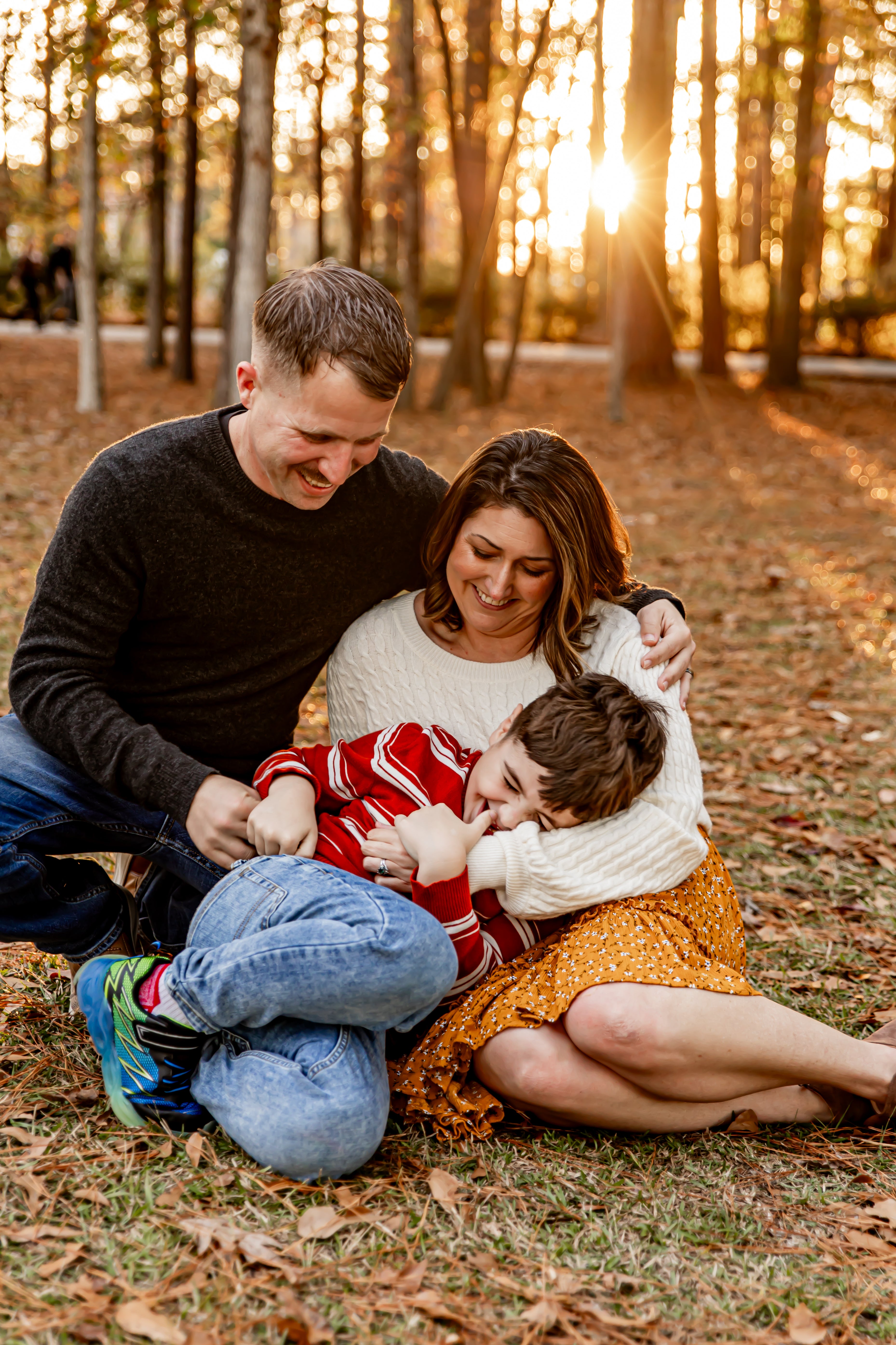 Family of three sitting on the ground in a park with trees and sunlight filtering through.