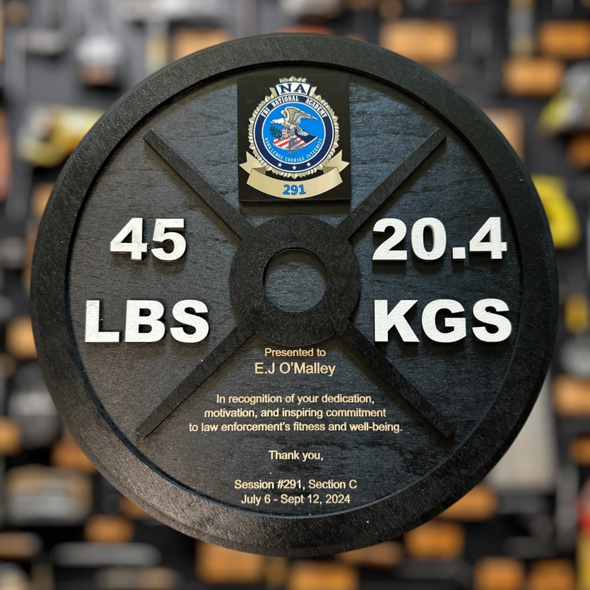 Weight plate with 45 lbs and 20.4 kgs markings, featuring a logo and text on a blurred background of gym equipment.