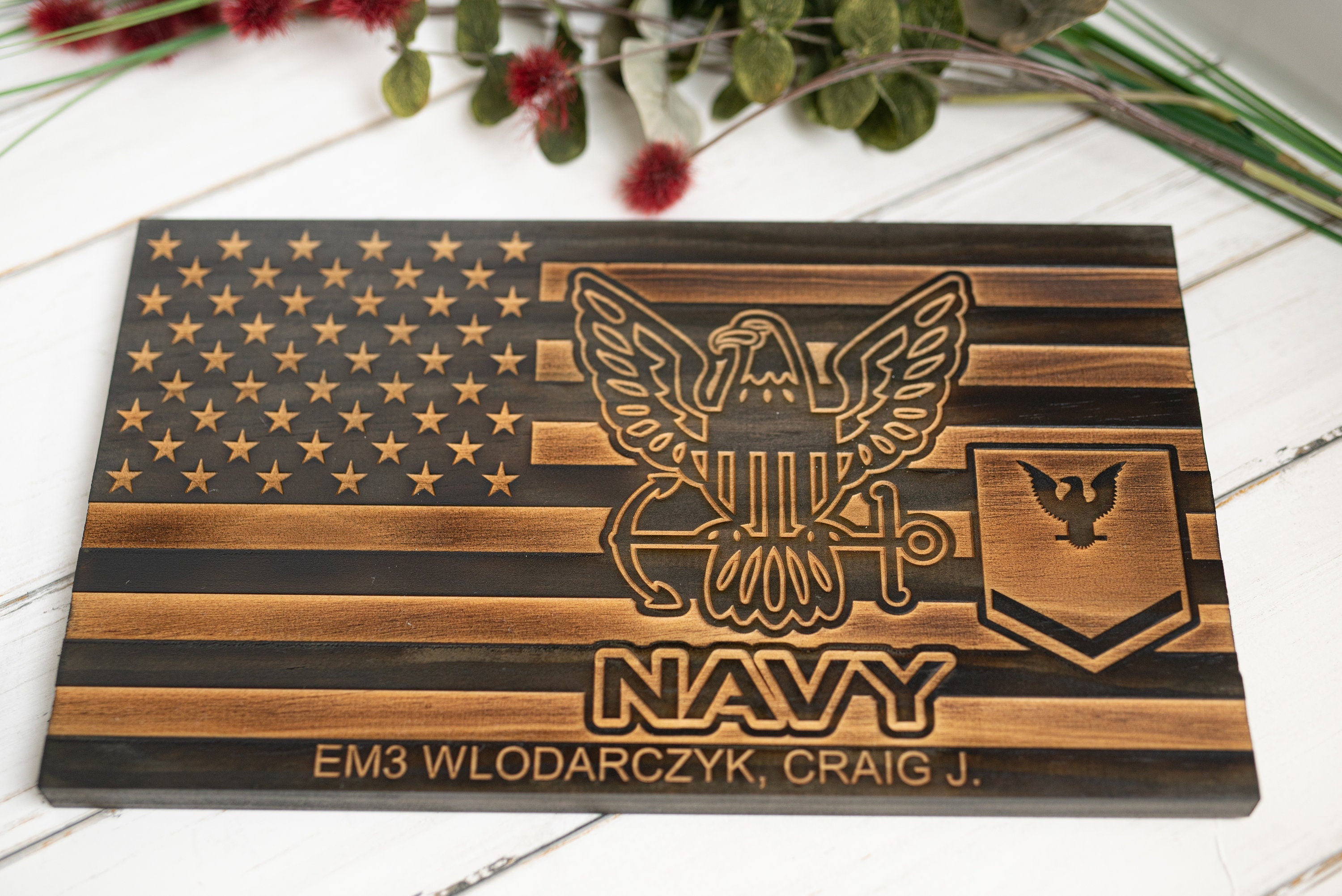 Ignite The Grain’s Navy Wooden American Flag Plaque for Veterans features an engraved flag, Navy emblem, “NAVY,” rank insignia, and the name EM3 Wlodarczyk, Craig J., set against a background with greenery and red berries.